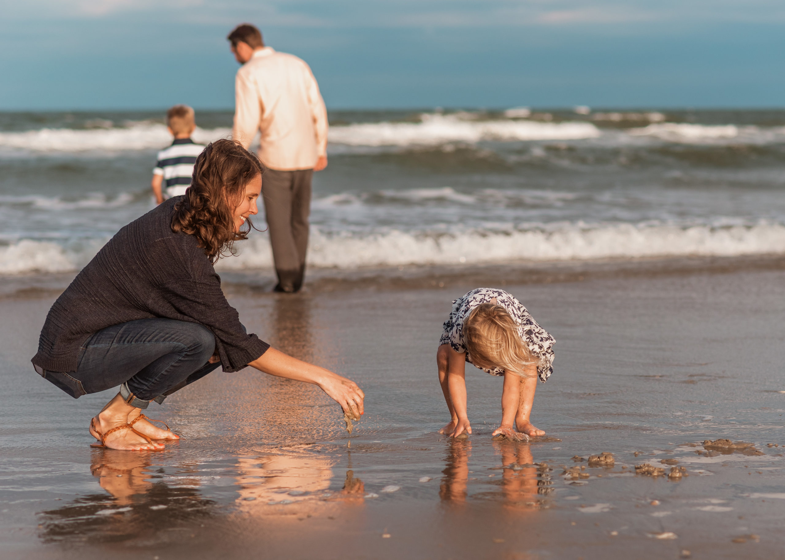 candid beach family shot by laura walter photography on the Outer Banks, mom and little girl playing in the sand while dad and son are in the ocean in the background